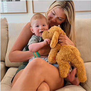 Smiling woman holding a happy baby who is playing with a brown stuffed dog on a beige couch.
