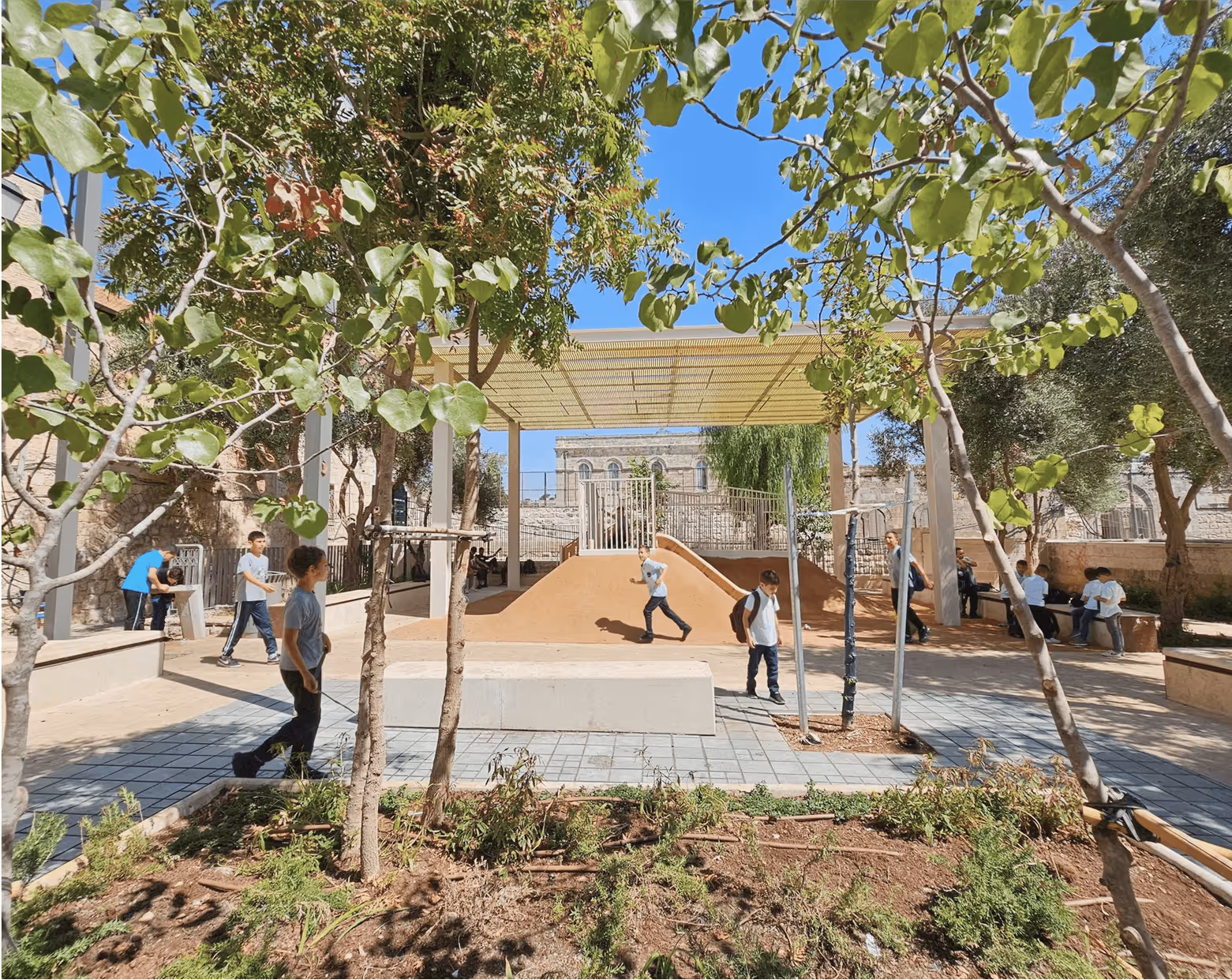 Children playing in a shaded outdoor park area with trees and playground equipment under a clear blue sky.