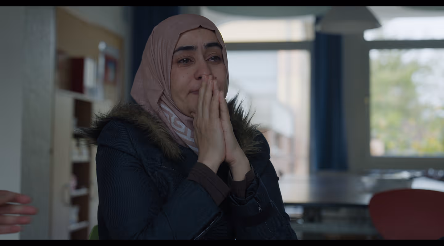 A still frame of the feature documentary Dancing Through: Woman wearing a pink hijab and dark coat with fur collar, crying and holding her hands to her face in an indoor setting.