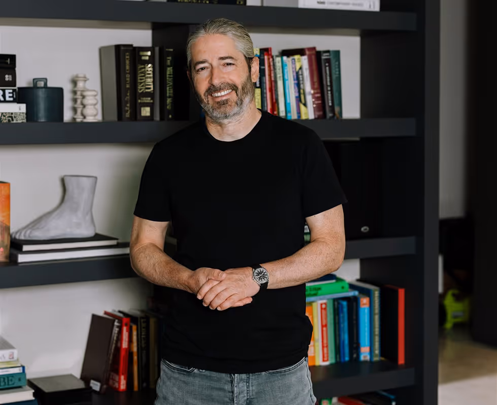 Ronnen Harary sitting cross-legged on a tan chair in front of bookshelves filled with books at his home in Toronto.