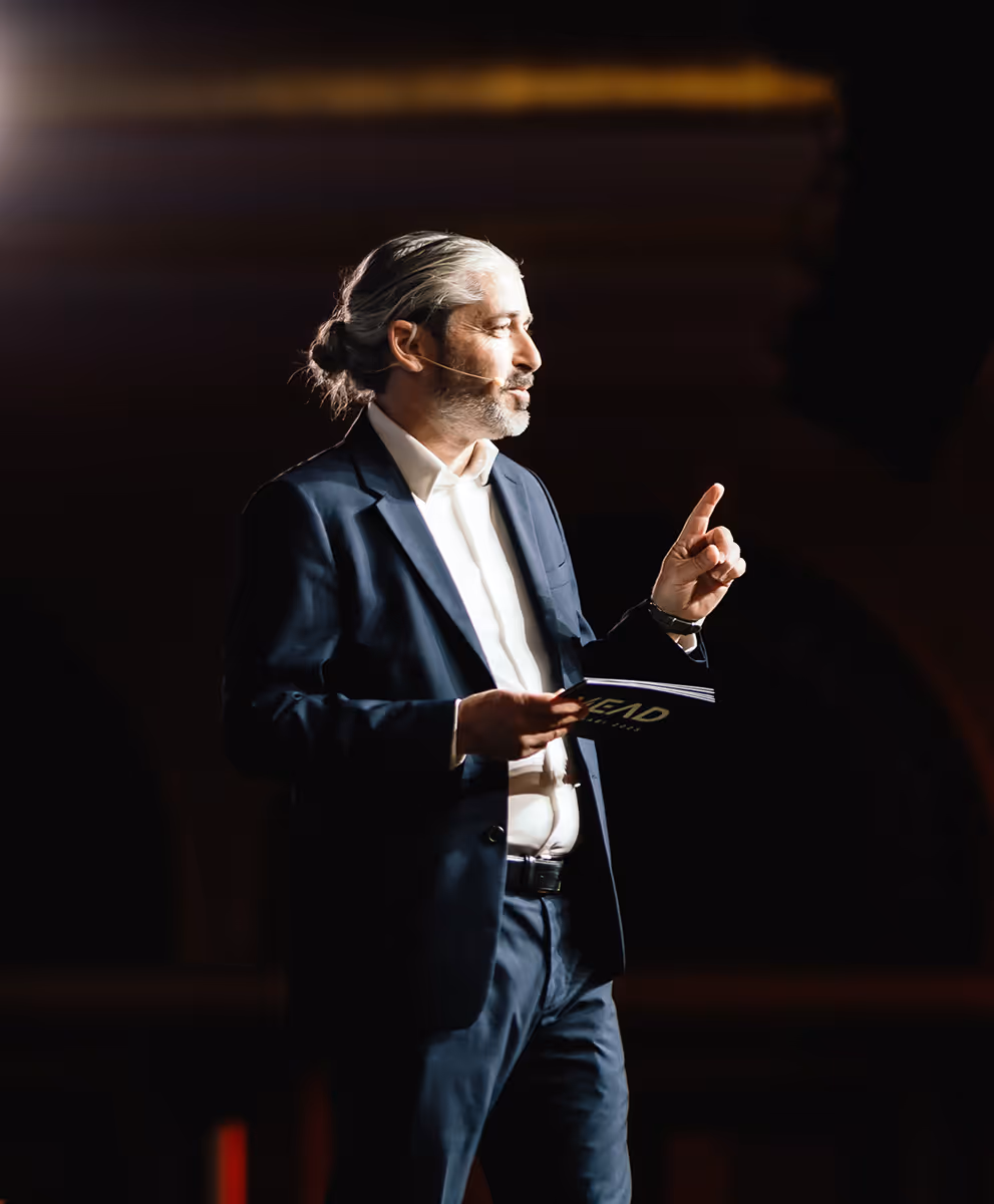 Ronnen Harary speaking on stage at the MEAD Summit, holding a MEAD program and gesturing to the audience