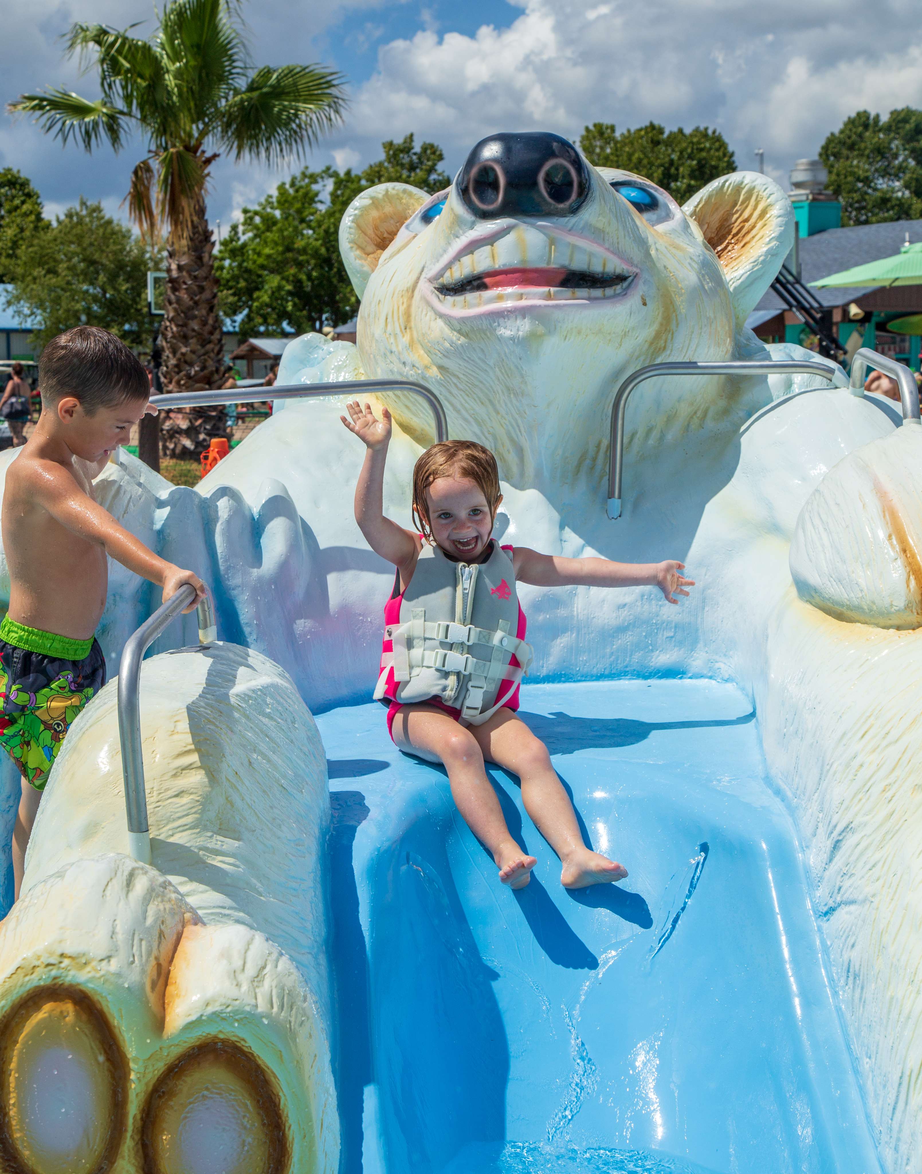 A young girl wearing a lifejacket smiles and waves from the top of a waterslide while a young boy waits his turn from the ladder. 