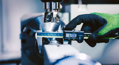 The image depicts a scene that appears to be from an industrial manufacturing or engineering setting. Here is a step-by-step description of the visible elements:

1. **Digital Caliper**: In the foreground, a digital caliper is being used to measure a cylindrical part that is mounted on what looks like a machining center. The caliper's display shows "62.08."

2. **Machining Center**: The machinery has a shiny metallic finish and consists of a spindle (not visible) and possibly various tools or fixtures attached to it. The part being measured is secured by clamps onto the worktable of the machine.