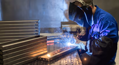 A person is welding metal indoors, creating sparks as they work. They are dressed in appropriate workwear for metalworking, highlighting their role as a technician or metalsmith. The scene emphasizes the hands-on nature of the task at hand.