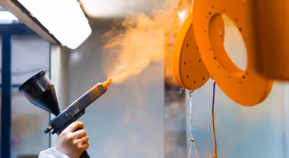 This is a photograph capturing the process of painting objects using an airbrush gun. The focus is on the act of spraying, where we see a fine mist of orange paint being dispensed from the nozzle of the gun.

The gun appears to be manual and is held by a person whose hand is not visible due to the protective cover or glove. The background reveals parts of what seems to be a paint booth, indicated by the partial view of the wall covered in white protective material which is typical for such environments to prevent paint splatters.