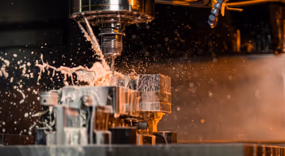 The image captures a dynamic industrial scene involving metalworking, specifically the process of machining or cutting metal using a high-precision machine tool, which appears to be a milling machine given the nature of the cut and the coolant being used.

The central focus is on the point where the cutting tool makes contact with a metallic workpiece that's secured in what looks like a vise. The water droplets and splashes in various directions suggest movement and action, indicating the machinery is actively working. The coolant is likely water, as it's a common choice for its ability to lubricate and protect the cutting edge from wear.