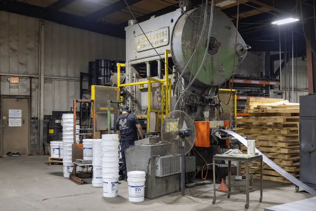 Worker operating a large Crown Boggs industrial stamping press in a factory, with buckets and stacked pallets nearby.