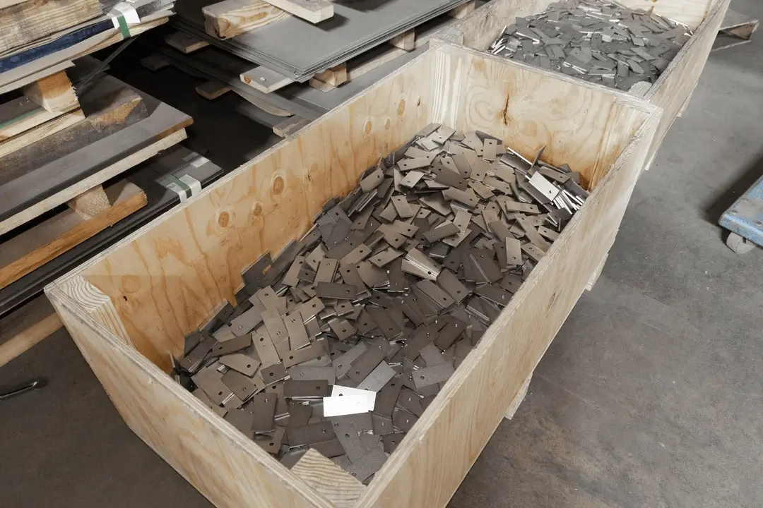 Wooden crate filled with stamped metal parts on a factory floor, with stacked metal sheets in the background.