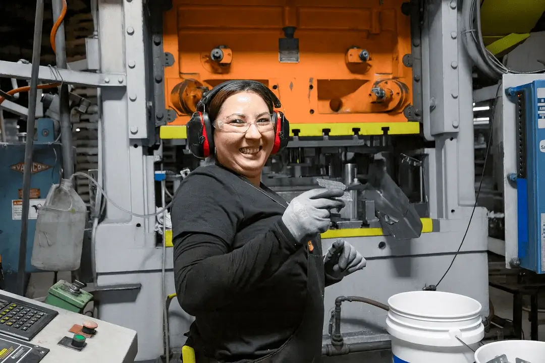 Worker wearing safety glasses and ear protection smiling and holding a metal part in an industrial stamping machine area.
