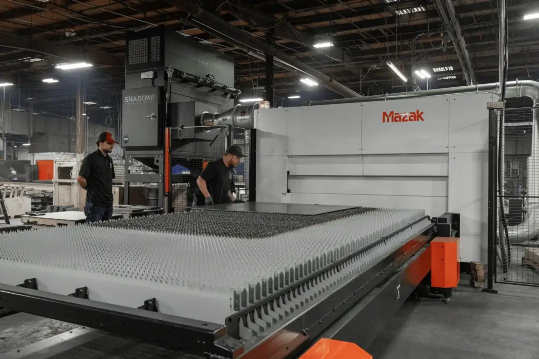 Two factory workers handle a large metal sheet near a fiber laser cutting machine in a manufacturing facility.