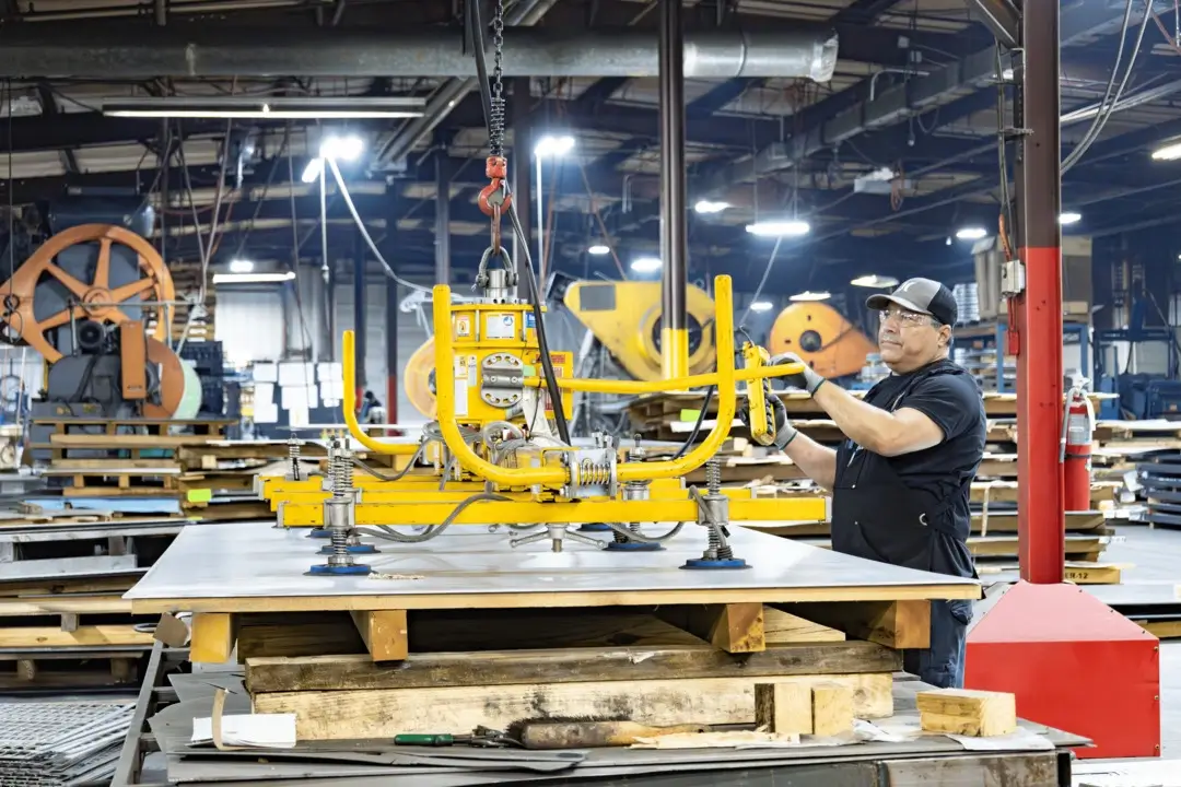 Factory worker operating a yellow vacuum lift to move a large sheet of metal on a worktable.
