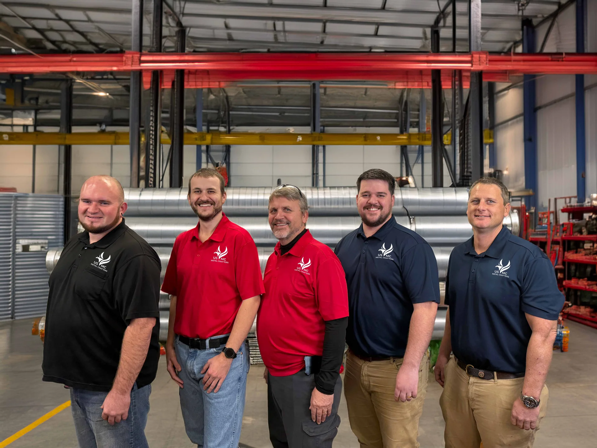 Five men in black, red, and navy polo shirts stand side by side in a workshop with industrial equipment behind them.