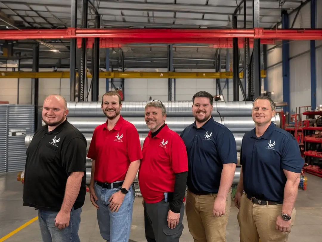 Five men in black, red, and navy polo shirts stand side by side in a workshop with industrial equipment behind them.