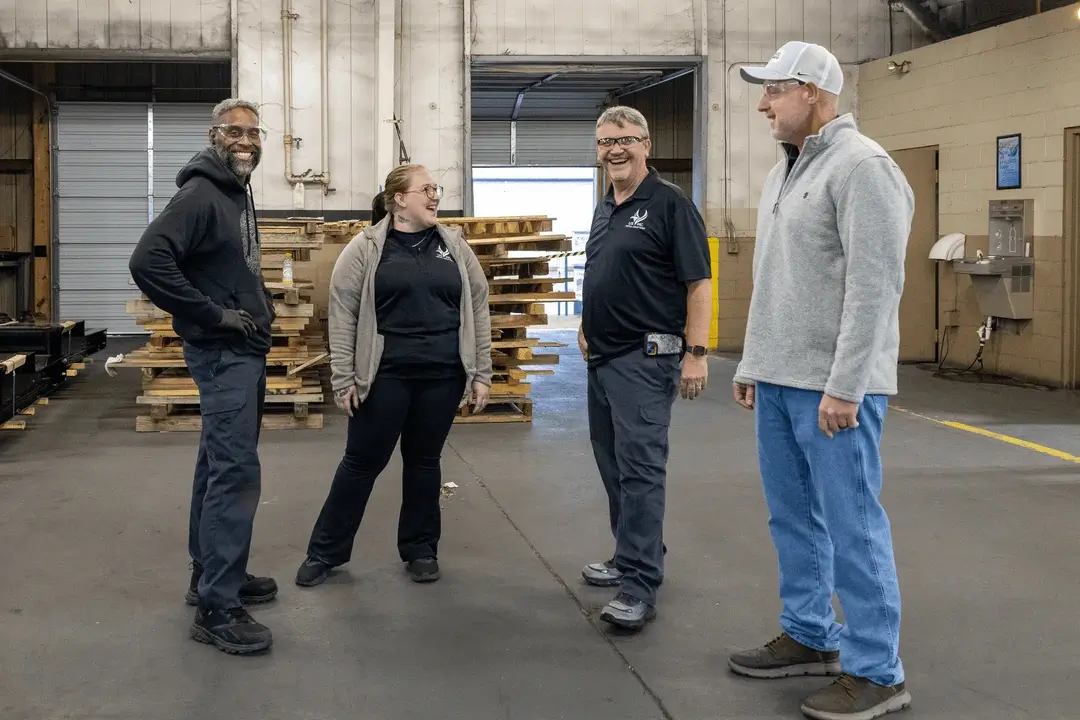 Four adults wearing safety glasses standing and smiling in a warehouse with stacked wooden pallets in the background.