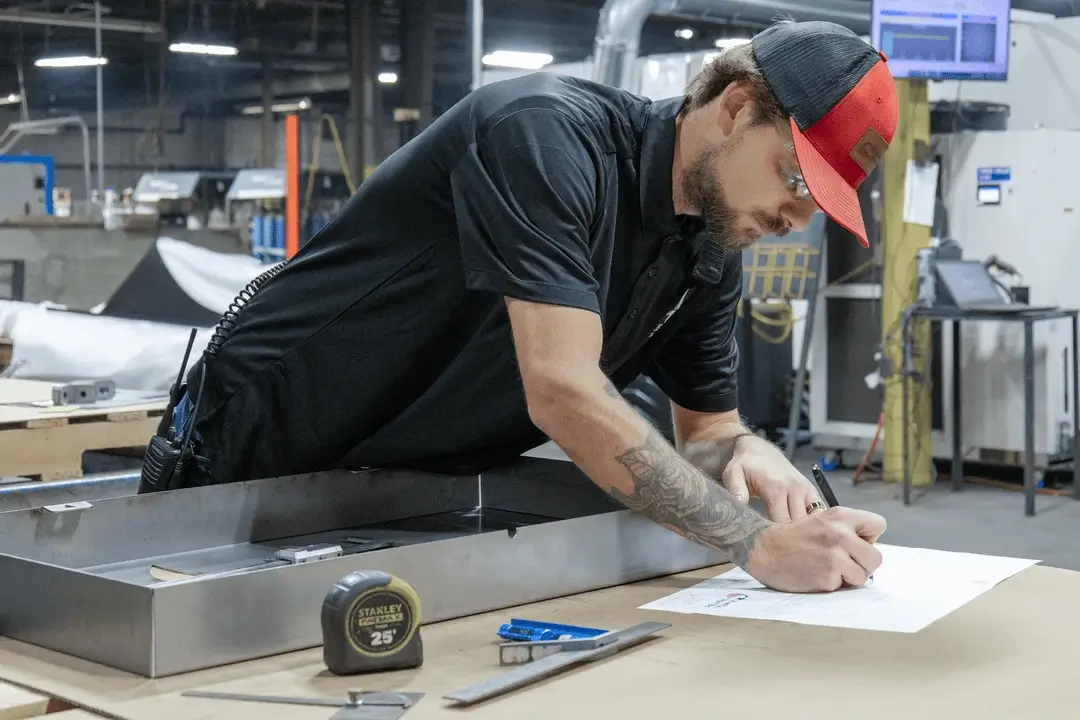 Worker in a red cap reviews a drawing and measures metal parts at a workbench in a manufacturing facility.