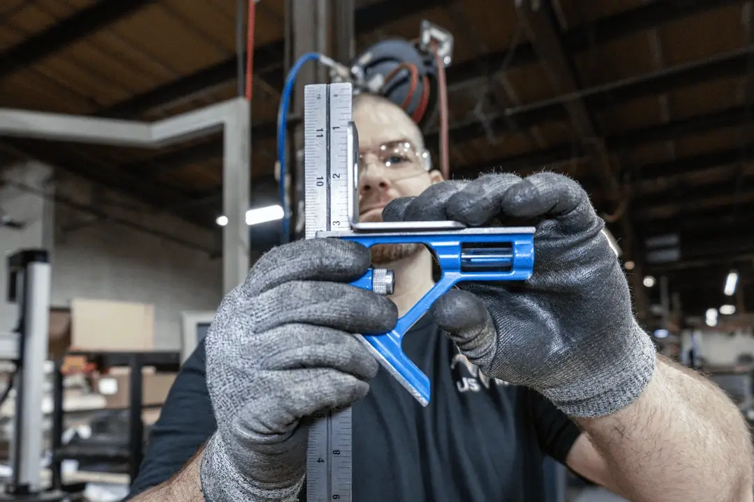 Man wearing safety gloves and glasses holding and measuring with a blue combination square tool in a workshop.