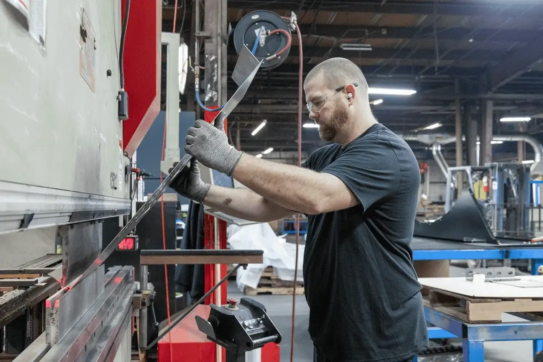 Man wearing safety glasses and gloves bending a metal sheet using an industrial press brake in a factory.