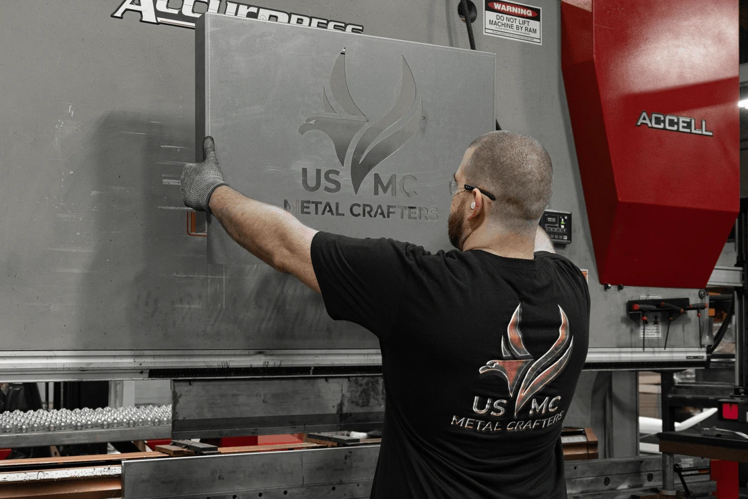 Worker wearing USMC Metal Crafters shirt handling a metal sheet with matching logo in front of a large Accell machine.