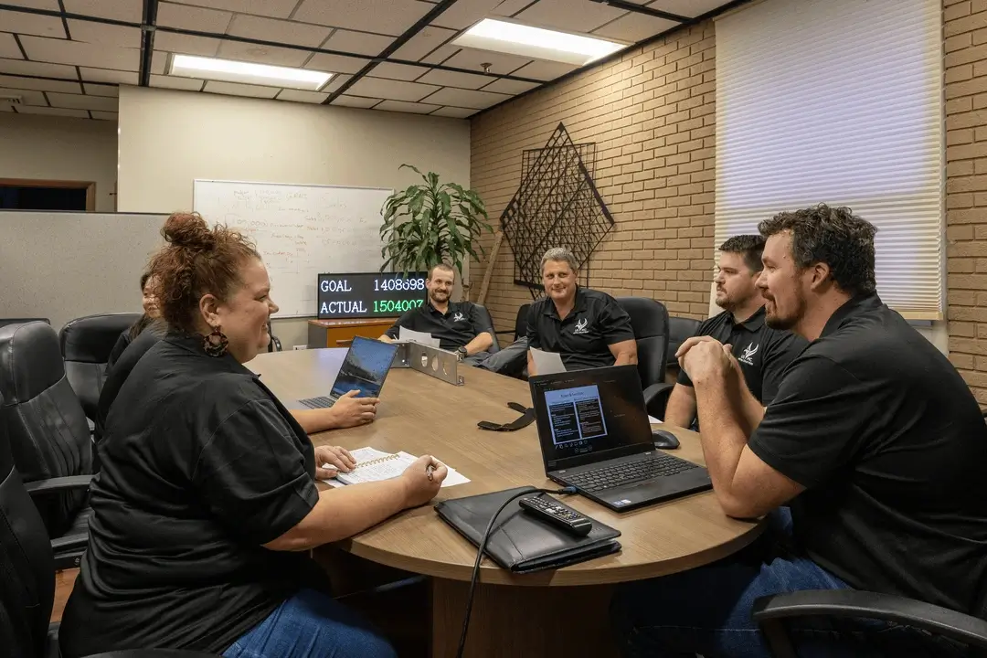 A group of six colleagues in black shirts seated around a conference table having a meeting with laptops and papers.