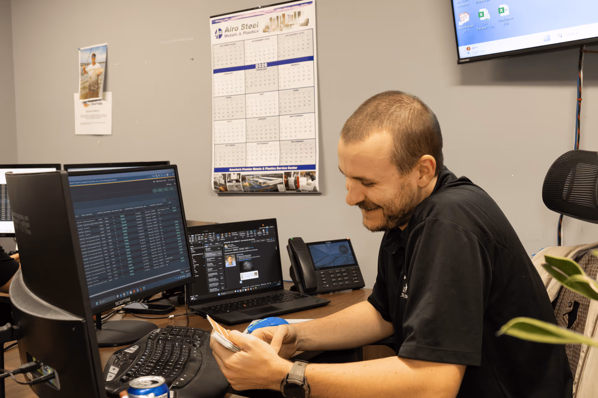 Man smiling while sitting at a desk with two computer screens, a keyboard, and a phone in an office.