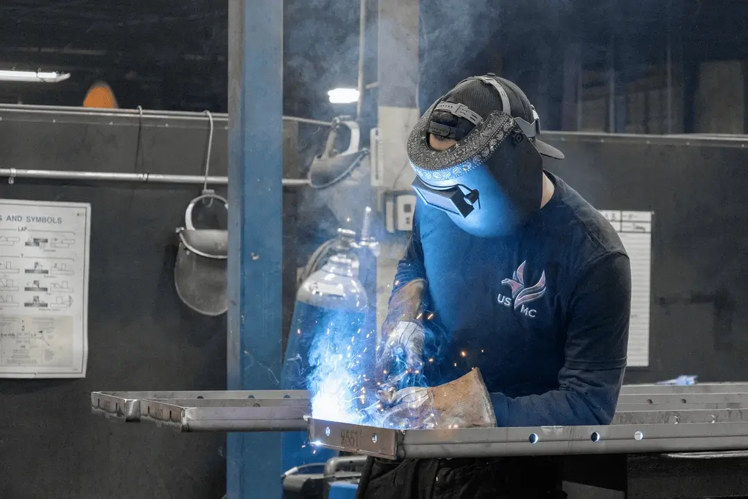 Welder wearing a protective helmet welds a metal frame, with sparks and smoke in a factory workshop.