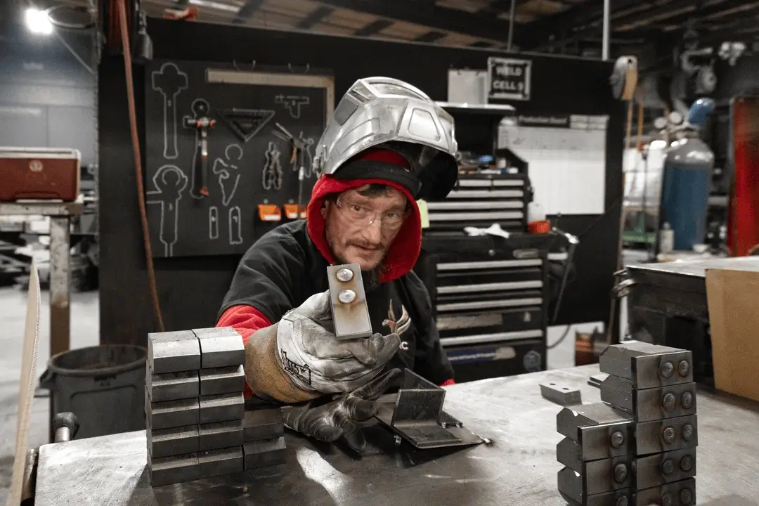 Worker wearing safety glasses and a welding helmet holds up a small metal part at a workbench in a fabrication shop.