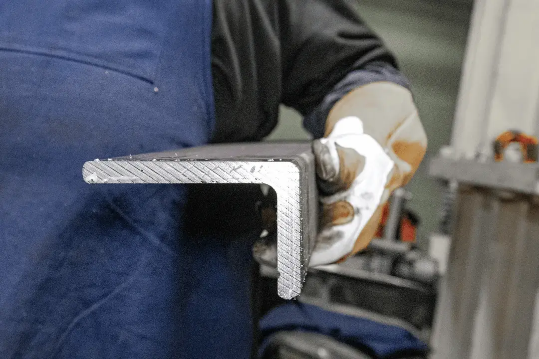 Gloved worker holds an L-shaped metal component, showing a close-up of the cut edge.
