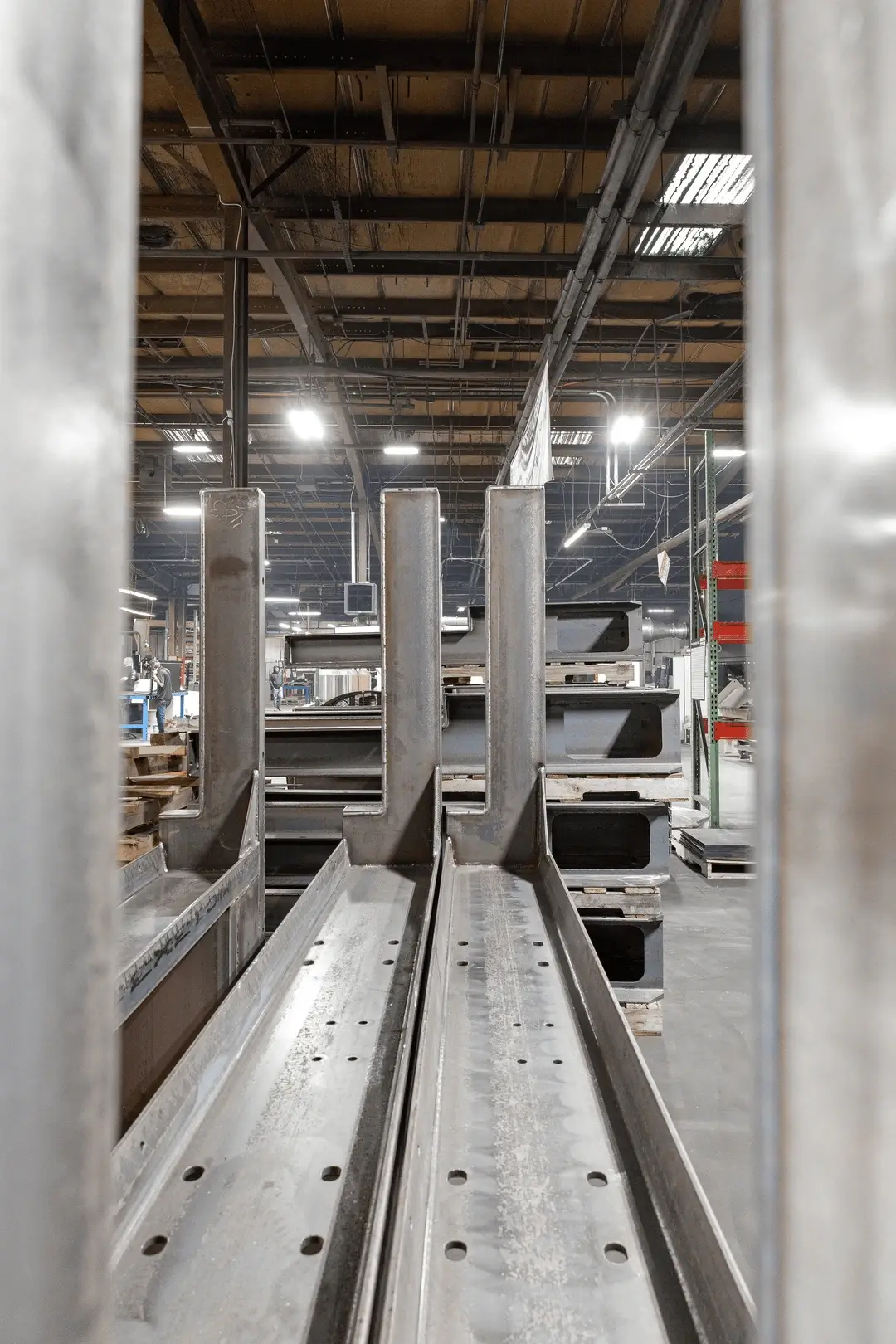Steel beams stacked in a manufacturing facility, viewed between two vertical supports with shop equipment in the background.