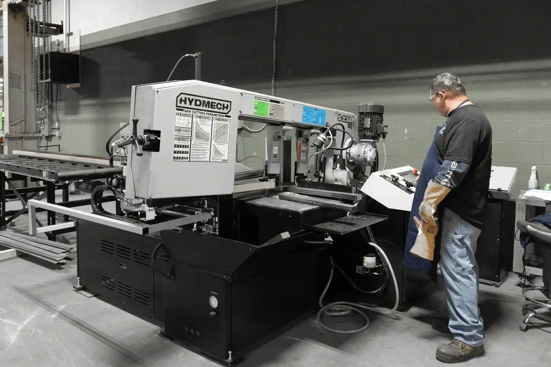 Man wearing protective gloves and apron operating a Hydmech S-23A metal cutting band saw machine in an industrial workshop.