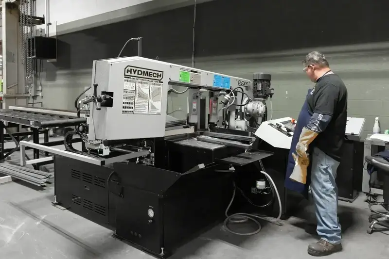 Man wearing protective gloves and apron operating a Hydmech S-23A metal cutting band saw machine in an industrial workshop.