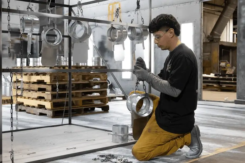 Factory worker wearing protective gloves and goggles kneeling while inspecting metal parts hanging from chains in an industrial workshop.