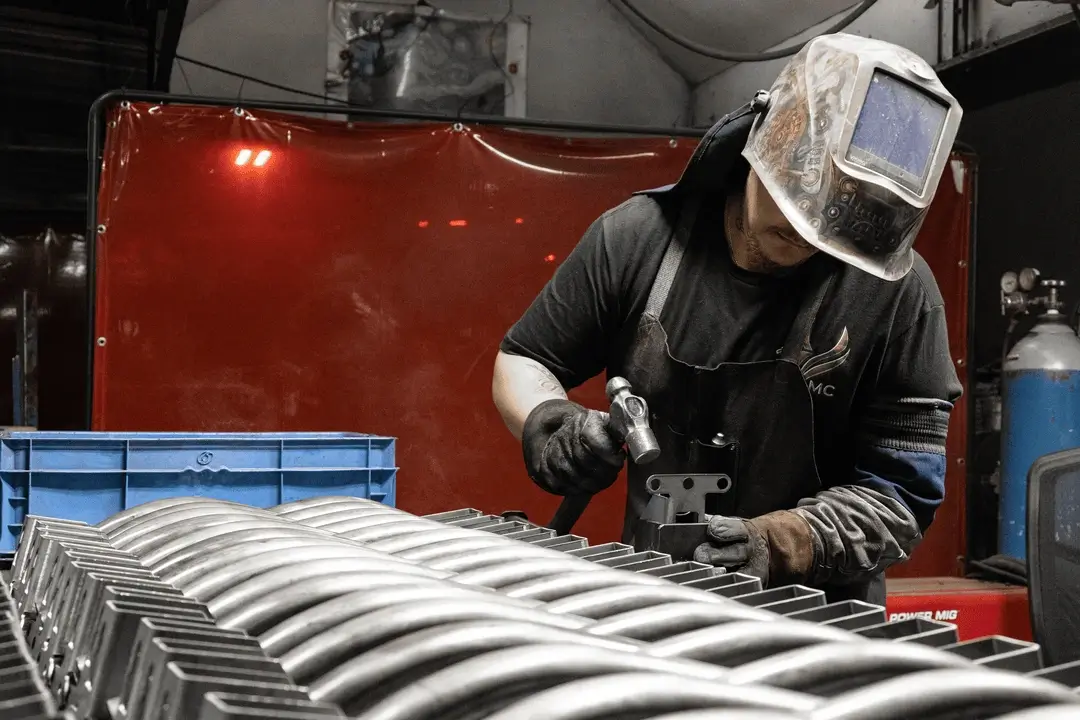 Worker wearing a welding helmet uses a hammer on metal parts at a workstation, with stacked components in front.