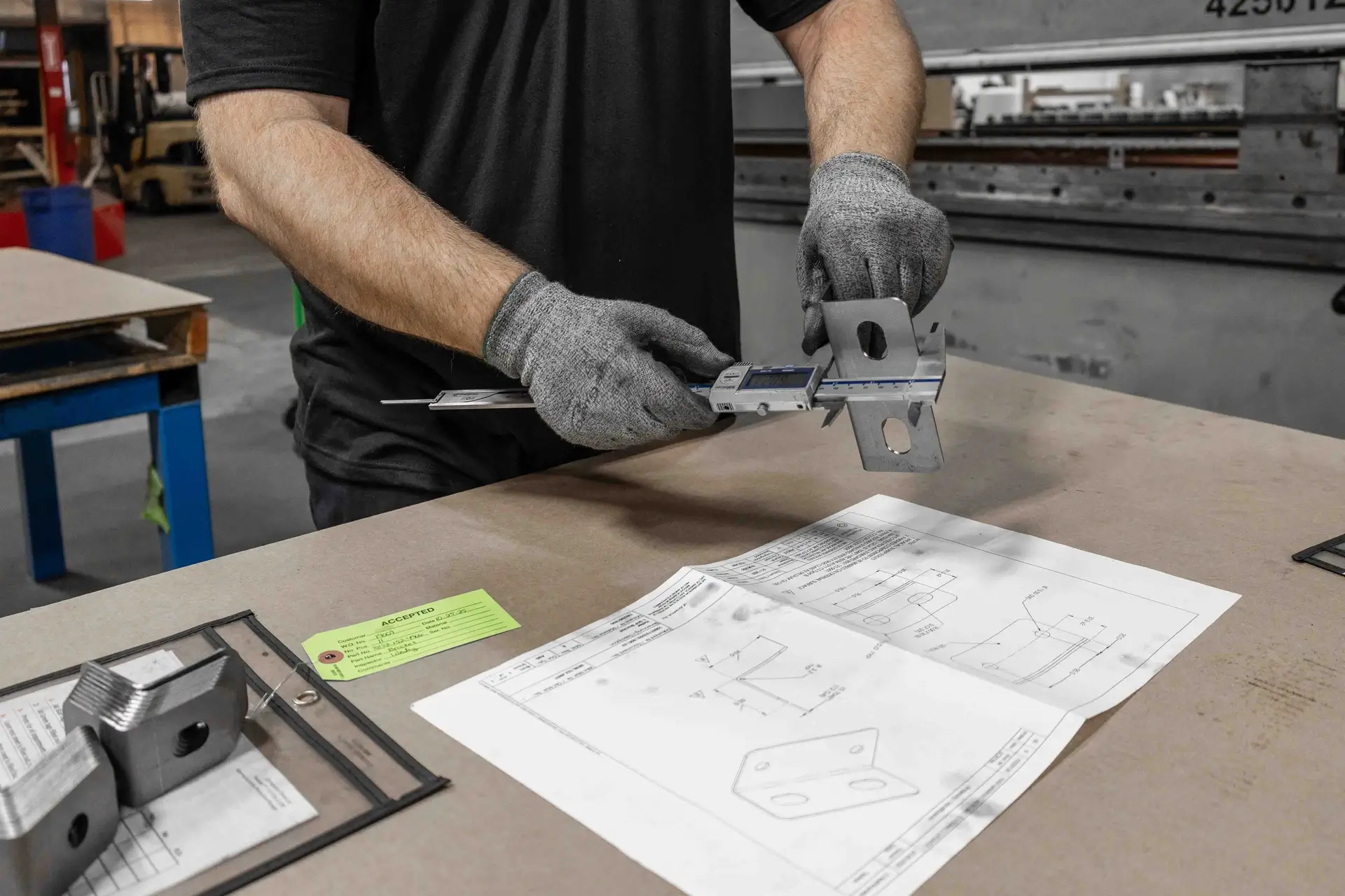 Person wearing gray gloves measuring a metal bracket with a digital caliper over a worktable.