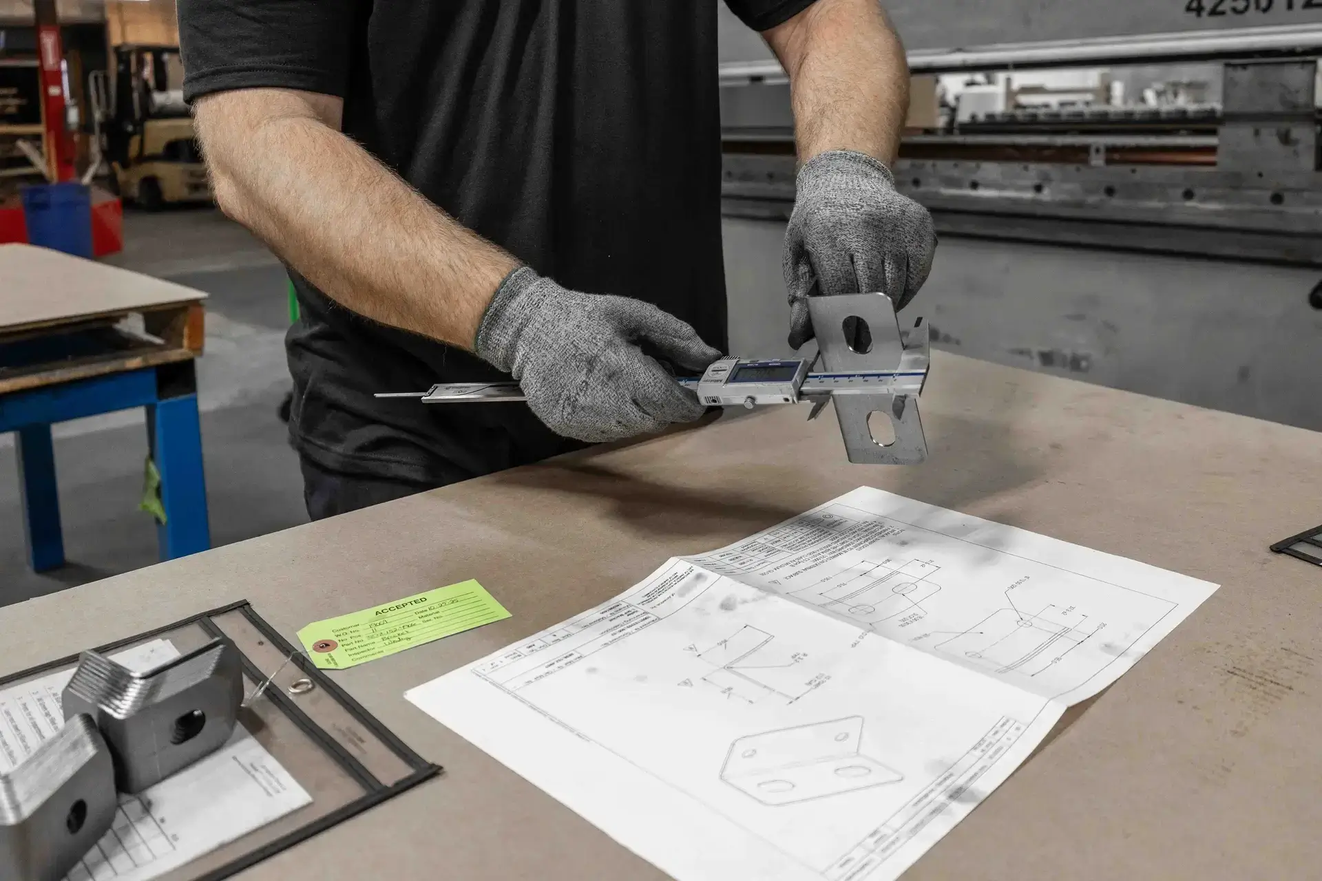 Person wearing gray gloves measuring a metal bracket with a digital caliper over a worktable.