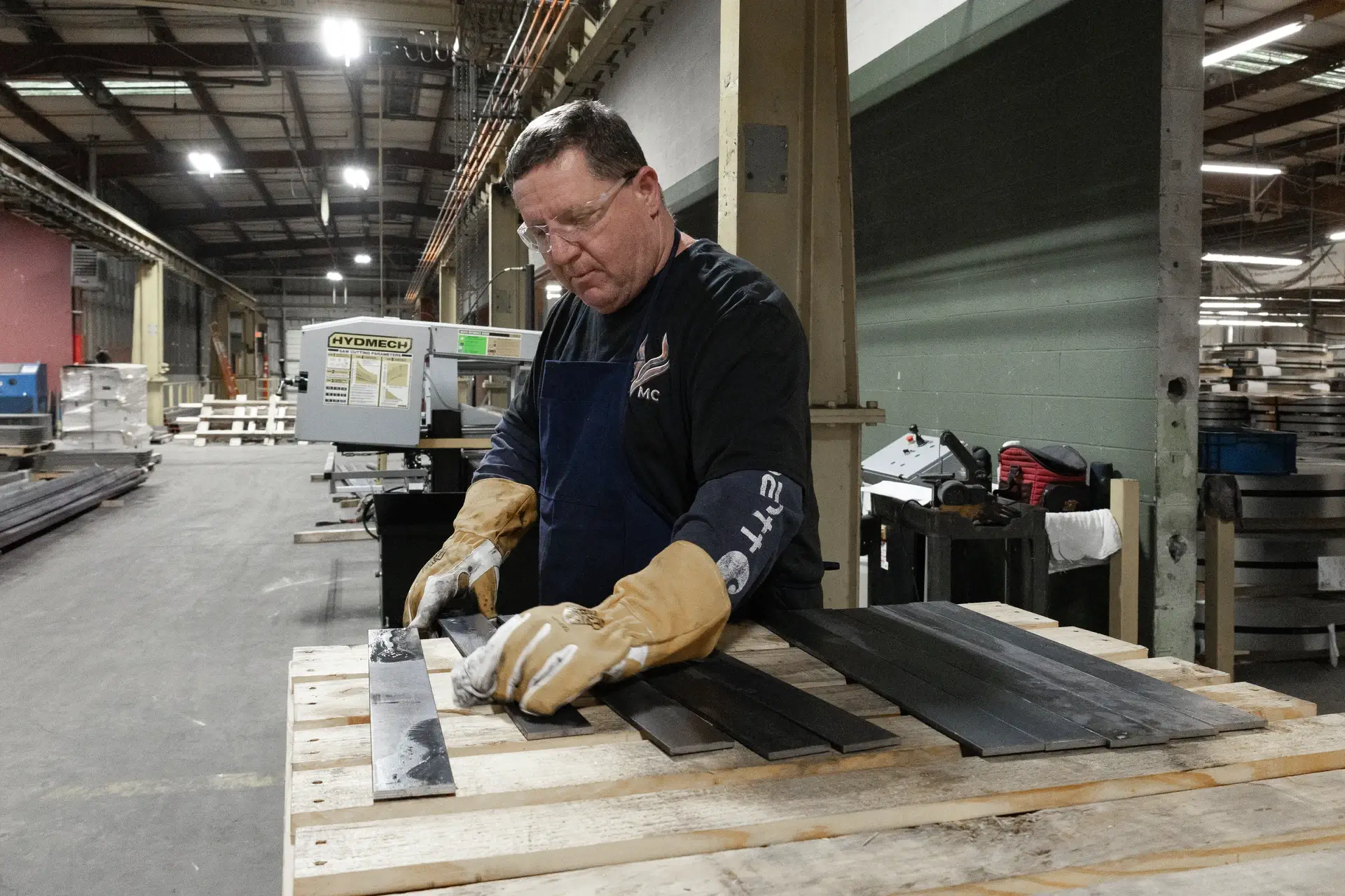 Man wearing gloves and safety glasses inspecting metal bars on a wooden pallet in an industrial workshop.
