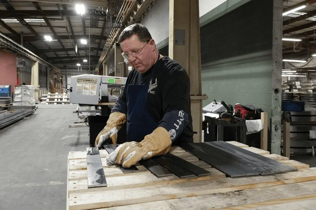 Man wearing gloves and safety glasses inspecting metal bars on a wooden pallet in an industrial workshop.