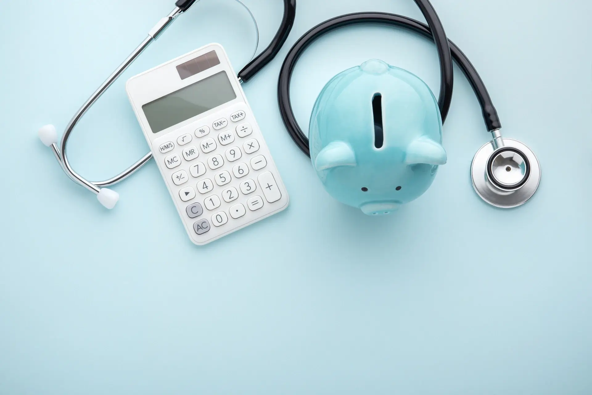 Blue piggy bank, white calculator, and black stethoscope on light blue background.