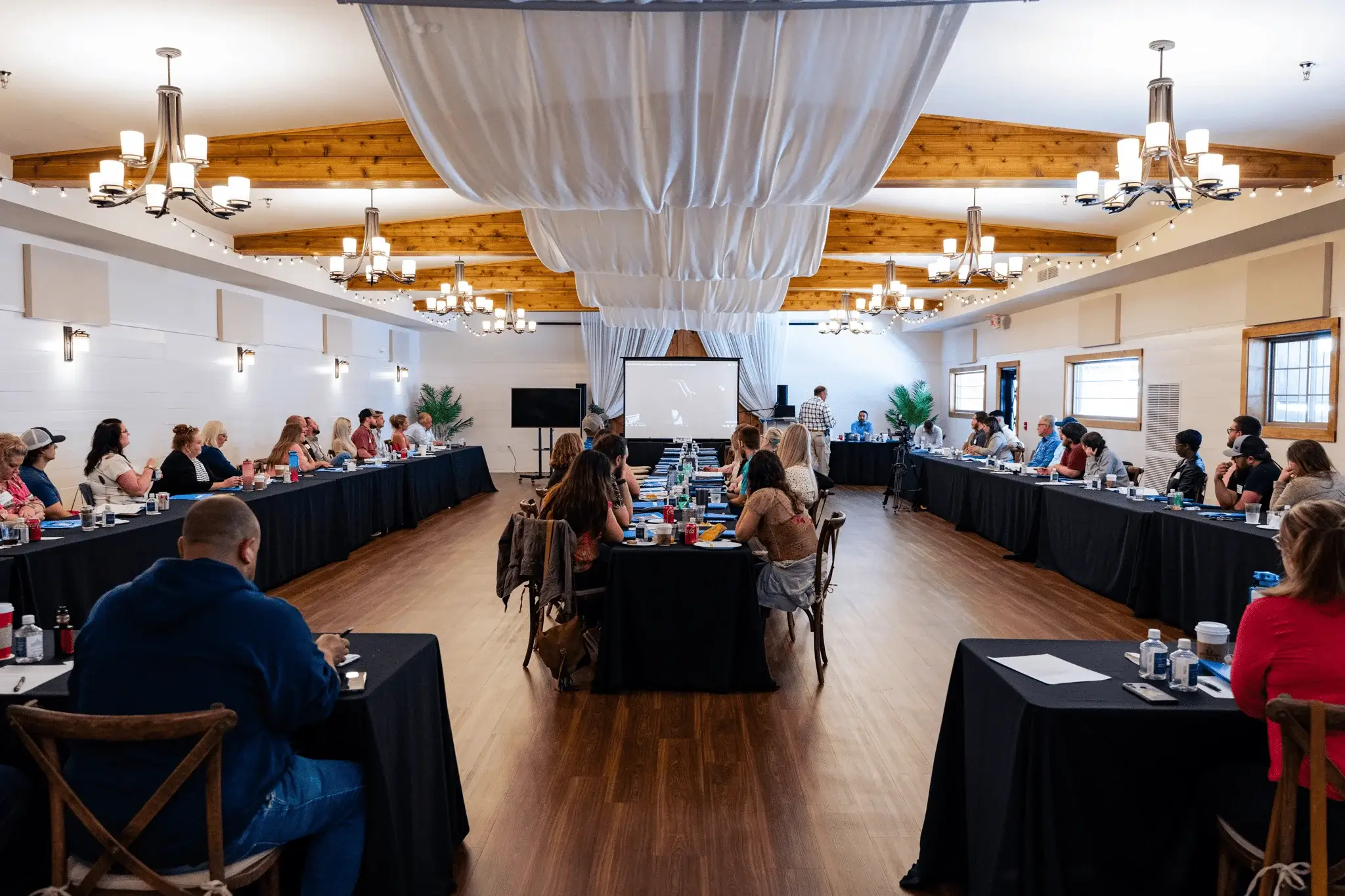 Large group of people seated around three long tables arranged in a U-shape in a spacious conference room with wooden beams and chandeliers.