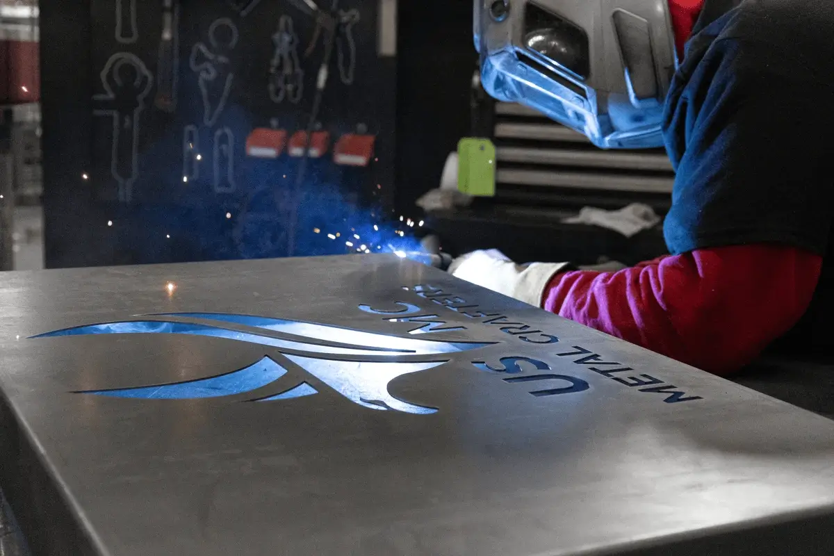 Welder in protective gear using a torch to cut or engrave a metal sheet with the inscription 'USMC METAL CRAFTSMAN'.
