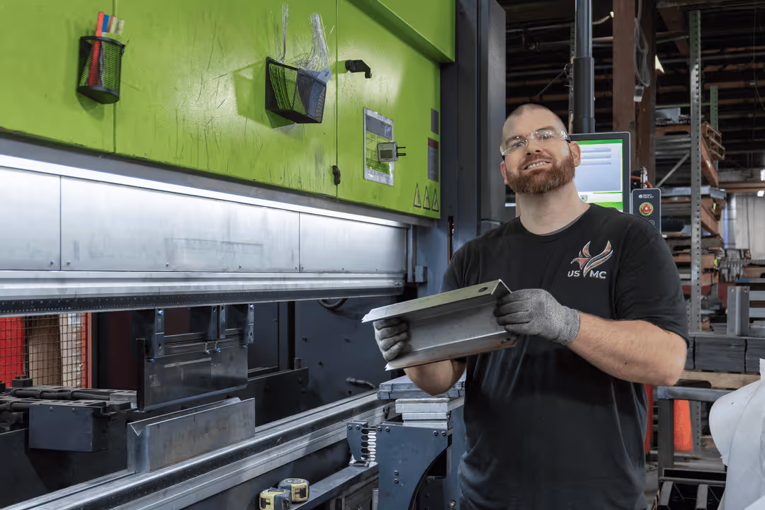 Man wearing safety glasses and gloves holding a metal piece in a factory with machinery in the background.