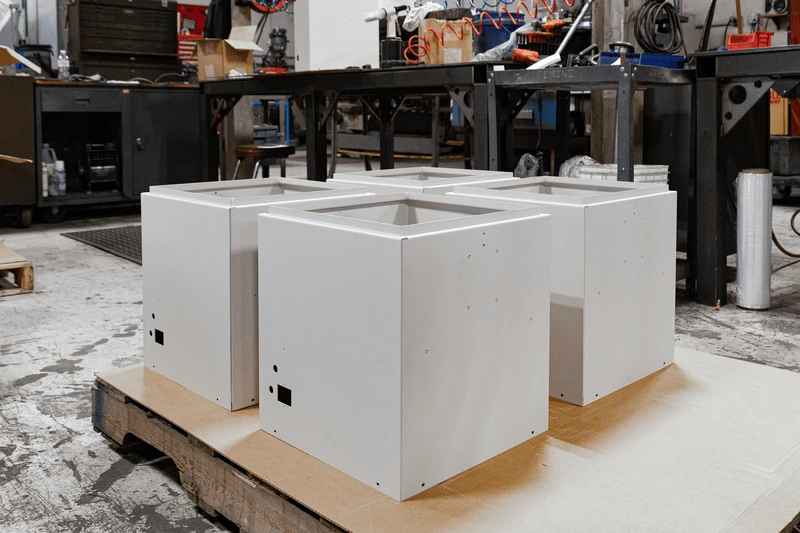 Four white metal enclosures placed on a wooden pallet in an industrial workshop.