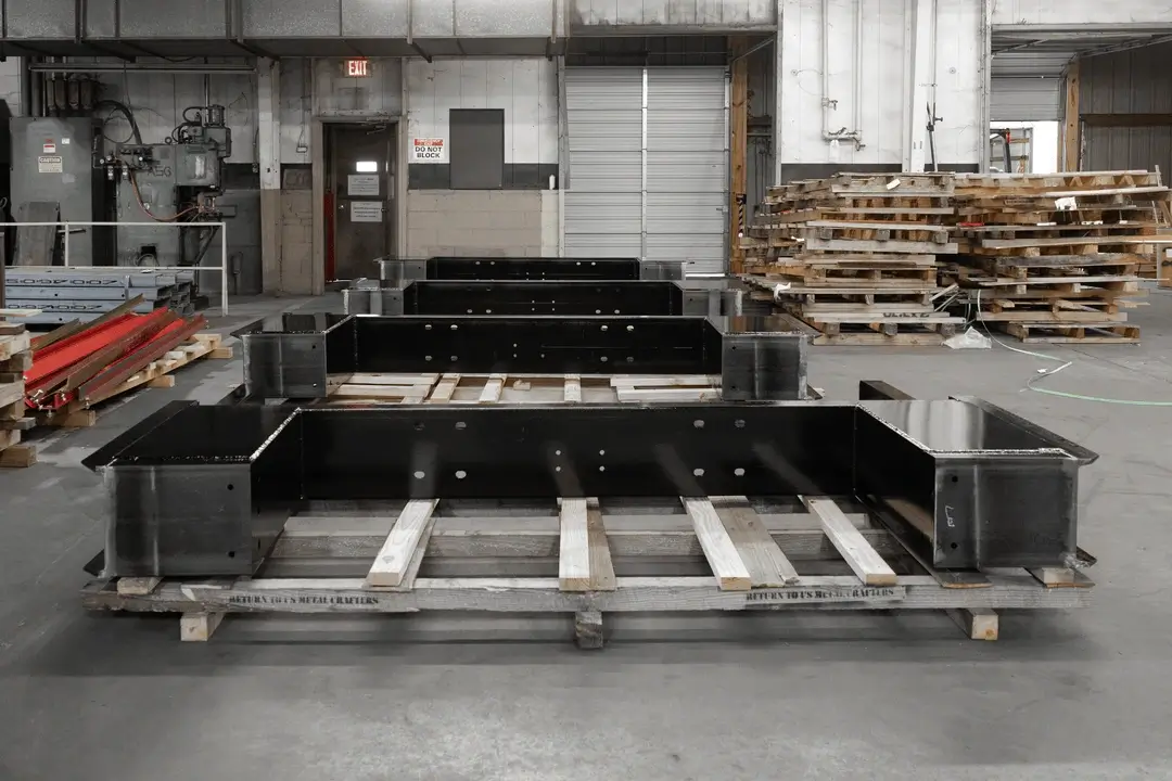 Industrial warehouse interior with black metal frames on wooden pallets and stacks of wooden pallets in the background.