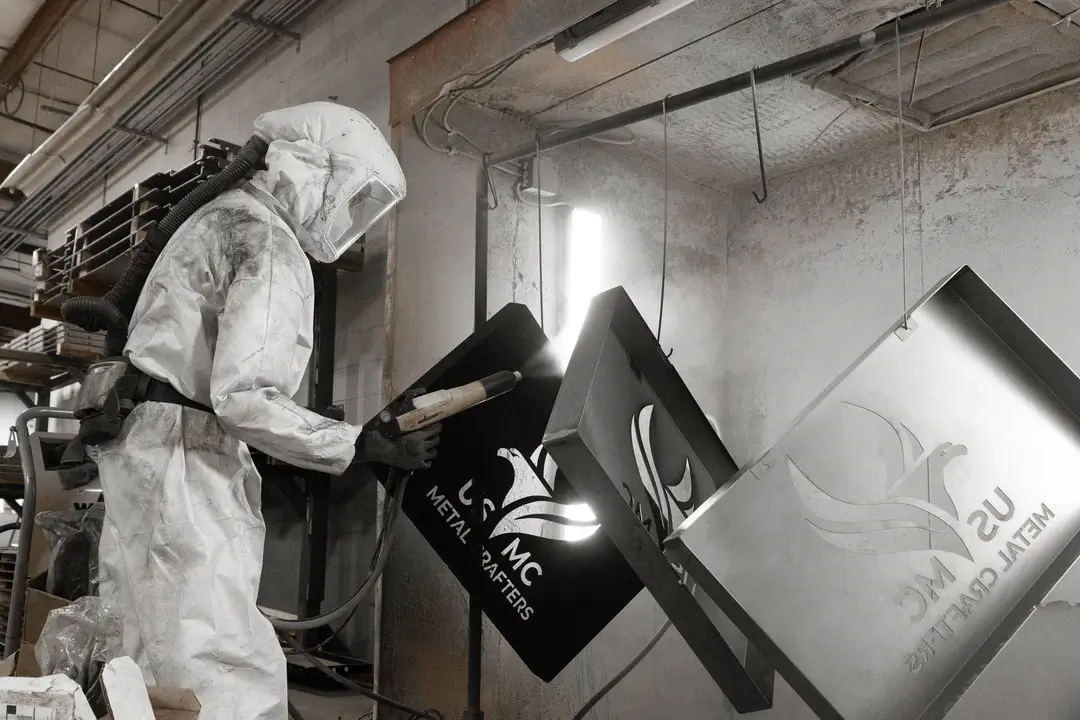 Worker in protective suit sandblasting metal signs with US MC Metal Crafters logo in workshop.