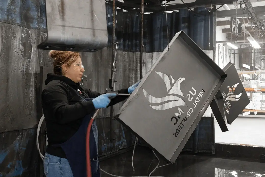 Woman wearing gloves and safety glasses working on a metal gate panel with a company logo in an industrial setting.