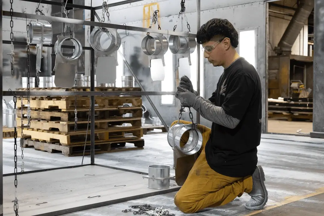 Worker wearing gloves and safety glasses kneeling while handling metal parts suspended on chains in an industrial warehouse.