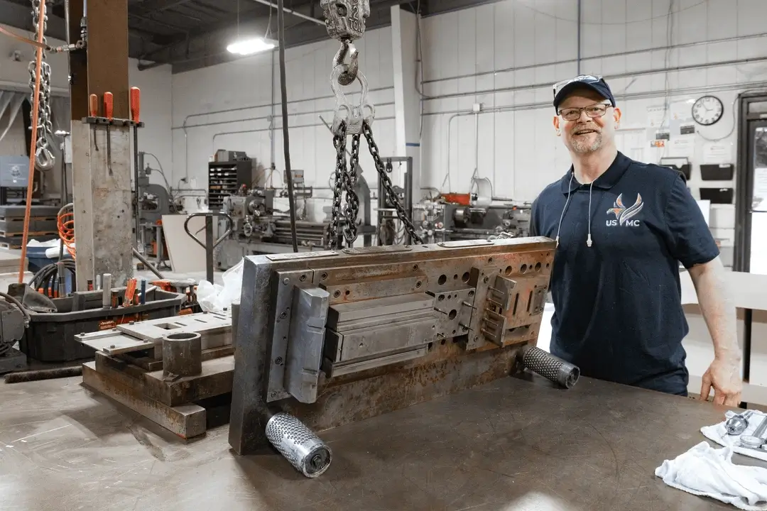 Smiling machinist stands beside a heavy metal die lifted by chains in an industrial workshop.