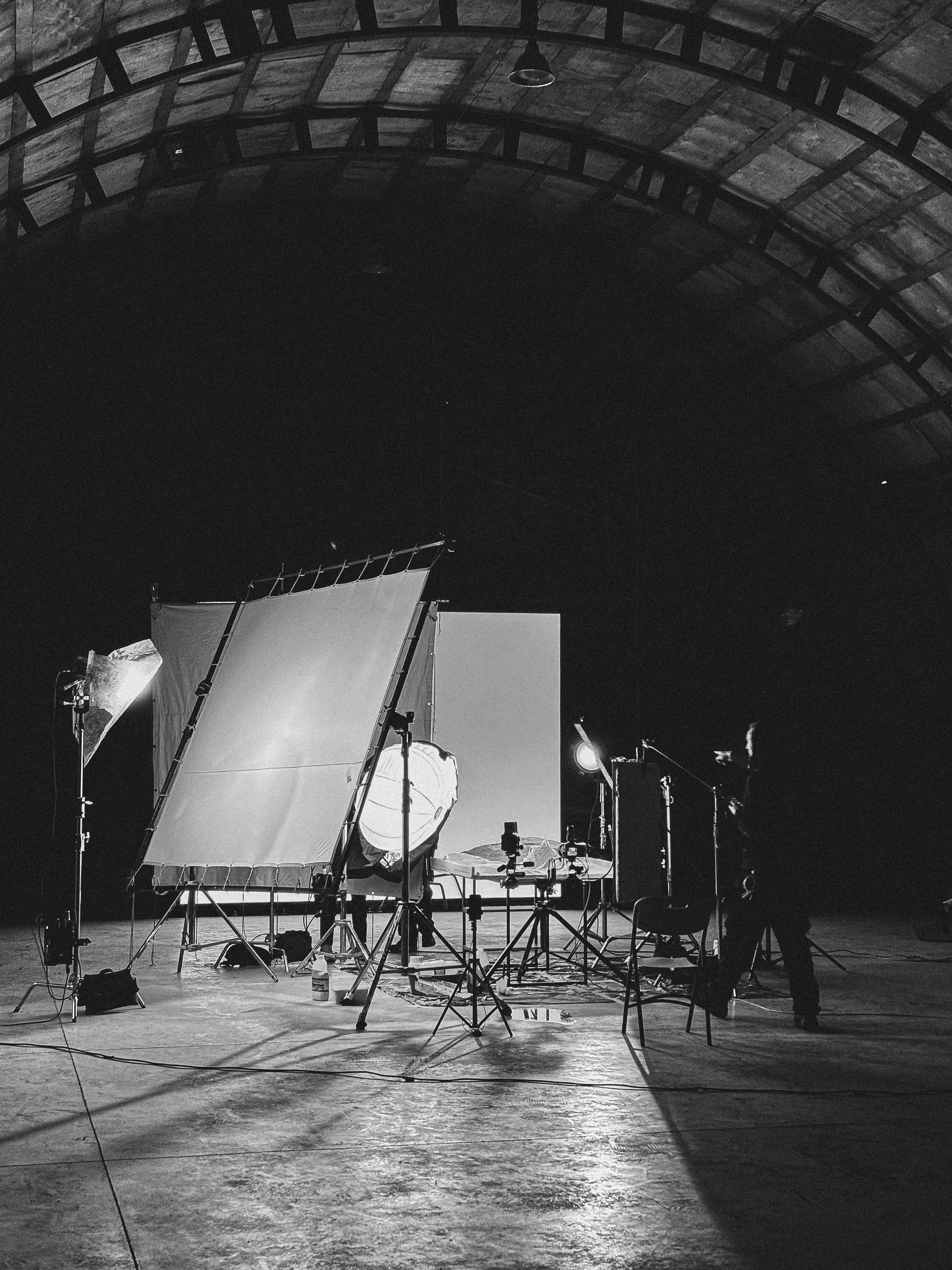 Black and white photo of a dimly lit film or photography studio with lighting equipment and a person adjusting the setup under a large arched ceiling.