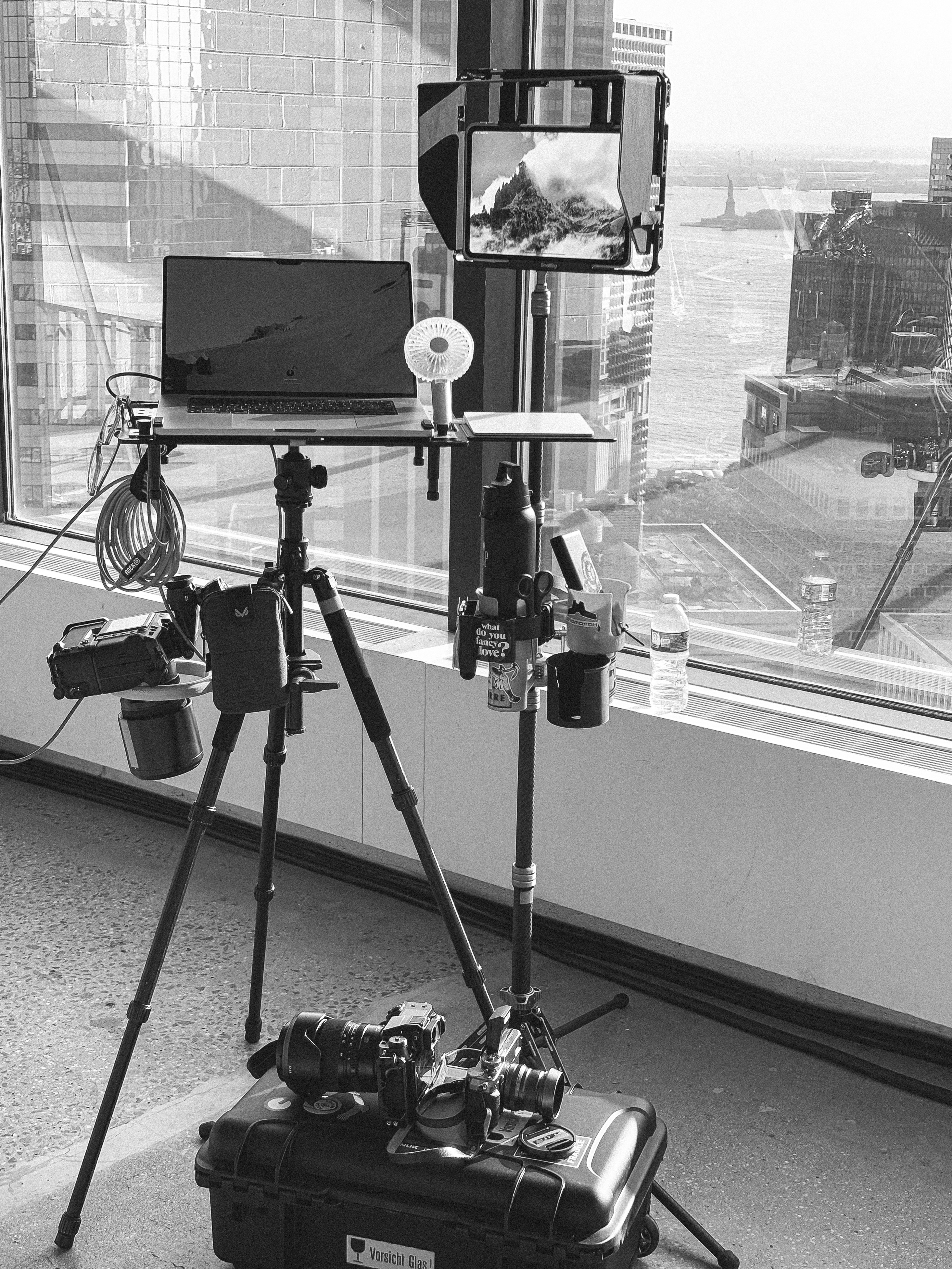 Black and white photo of a photography setup with a laptop on a tripod, a tablet mounted nearby, cameras on a case, and a cityscape with Statue of Liberty visible through large windows.