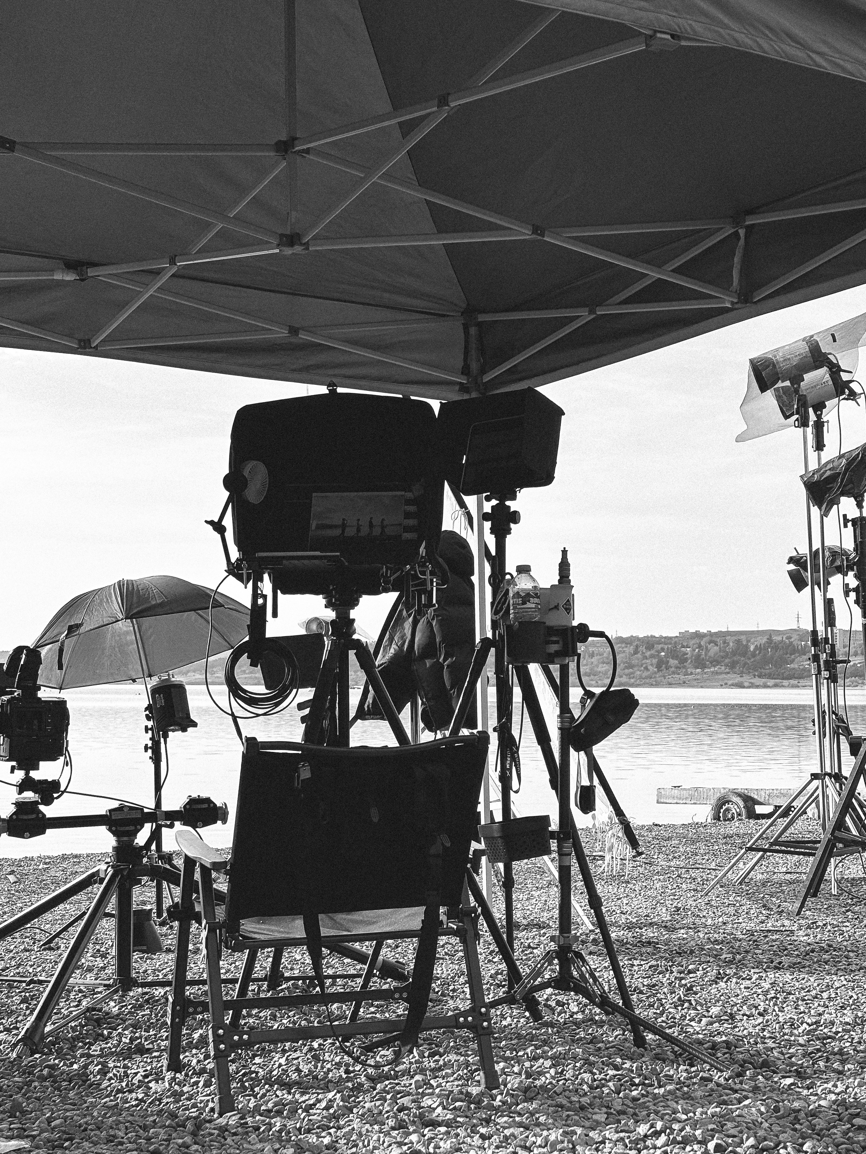 Black and white photo of film production equipment, lighting, and a director's chair set up on a rocky shore by a calm body of water.