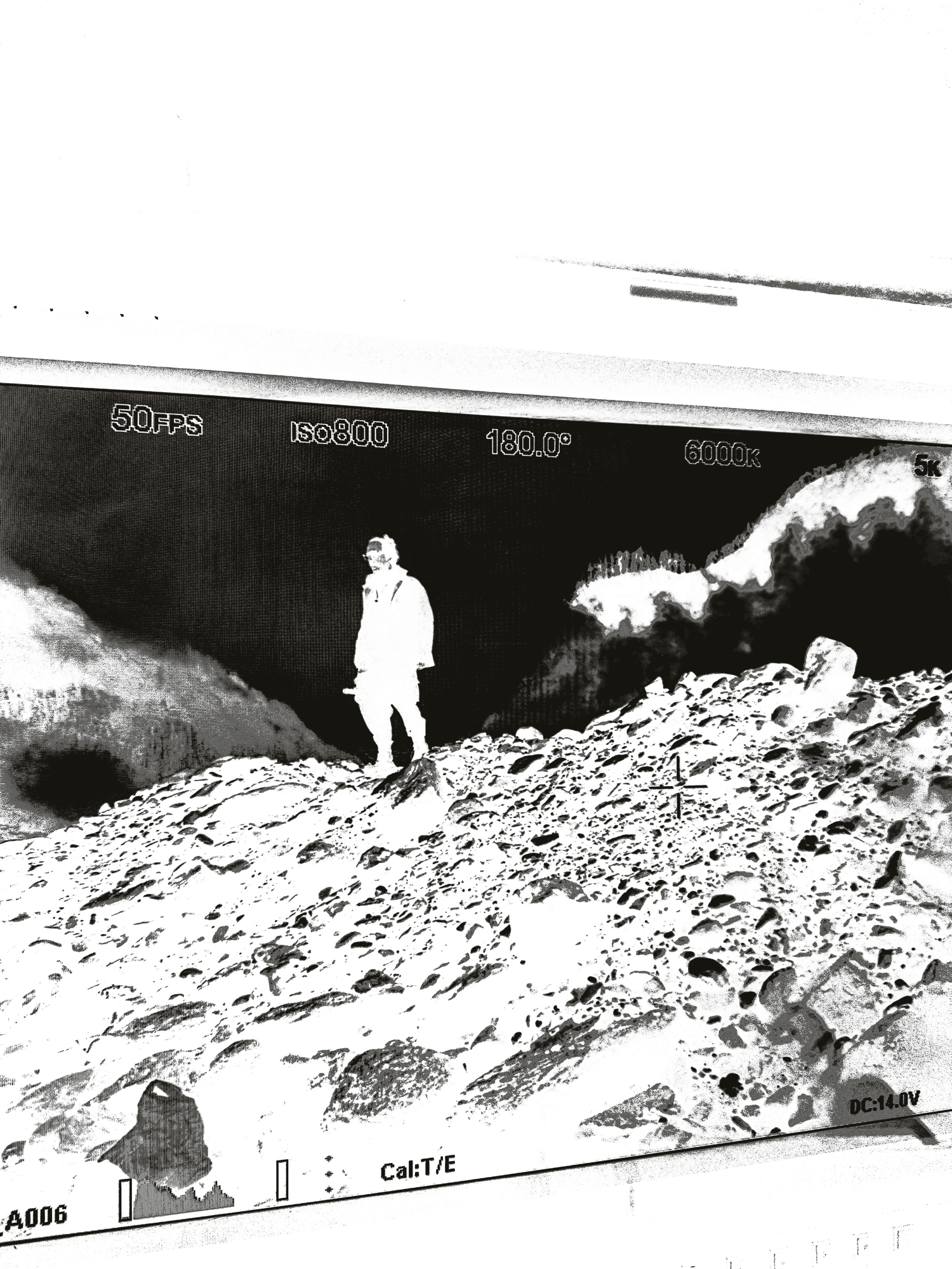 Camera screen showing a person standing on rocky terrain with mountains in the background and camera settings displayed.